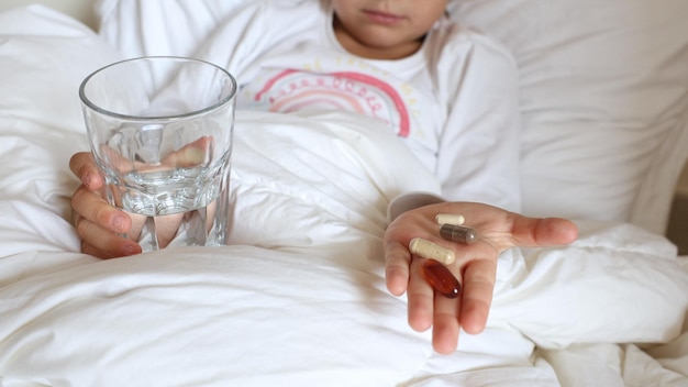 Nahaufnahme von Kinderhänden in der Handfläche, die ein Glas Wasser und medizinische Pillen halten. Krankes Kind liegt im Bett und ist bereit zur Einnahme