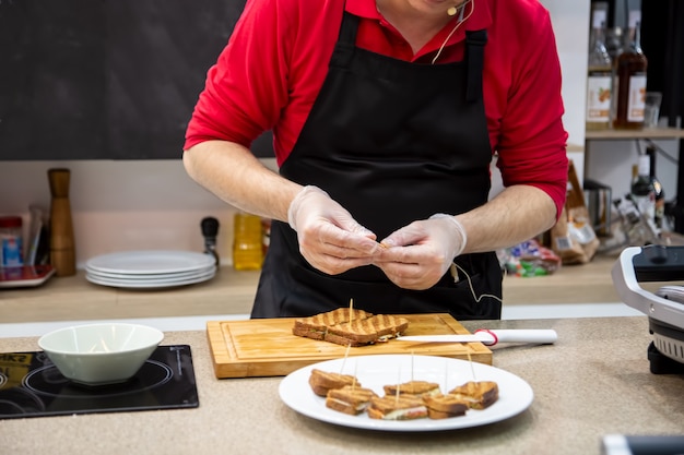 Nahaufnahme von Koch Mann in Gummihandschuhen bereiten Snacks gegrillte Sandwiches vor. Weichzeichner, Hintergrund ist Küche in Unschärfe