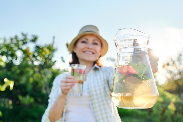 Nahaufnahme von Krug und Glas mit natürlichem Kräuterminze-Erdbeergetränk in den Händen der Frau. Sommerkalte Erfrischungsgetränke, gesunde hausgemachte Vitamingetränke