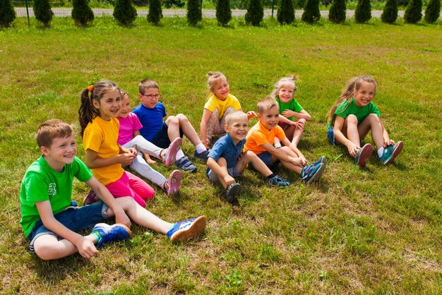 Nahaufnahme von lächelnden Kindern unterschiedlichen Alters in bunten T-Shirts, die auf einem grünen Rasen sitzen. Teilnehmer des Sommerfamiliencamps nach der Schule sitzen lächelnd und lachend auf einem Gras