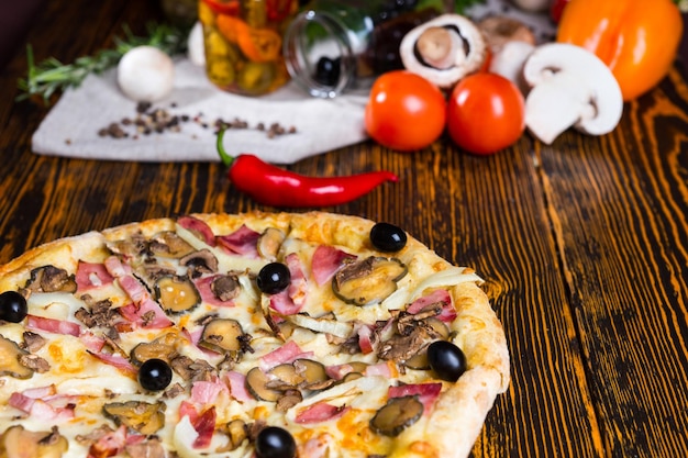 Nahaufnahme von leckerer Pizza mit Schinken, Zwiebeln, Oliven auf Holztisch, Tomaten, Pilzen und anderem Gemüse im Hintergrund