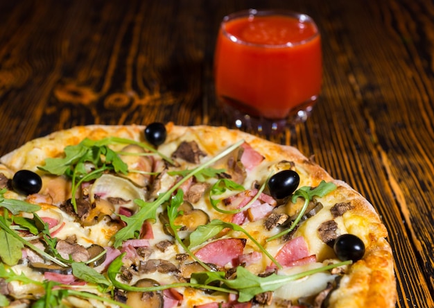 Nahaufnahme von leckerer Pizza mit verschiedenen Belägen und Käse auf Holztisch in der Nähe eines Glases mit Tomatensaft