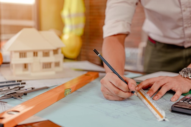 Nahaufnahme von männlichen Architektenhänden, die bei Sonnenuntergang ein Modellhaus auf dem Schreibtisch messen und herstellen. Ingenieur, Ingenieurwesen, Architektur, Design, Planung, Berufskonzept.