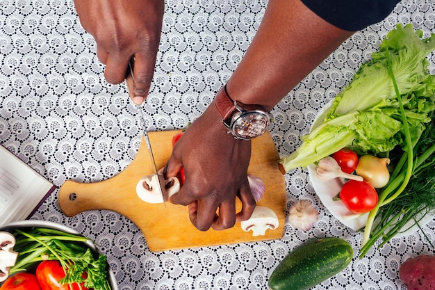 Nahaufnahme von Mannhänden Afroamerikaner schneidet Gemüse braten Salatpfeffer, Pilze, Tomate im Küchenrezeptbuch auf dem Tisch. veganes gesundes Essen