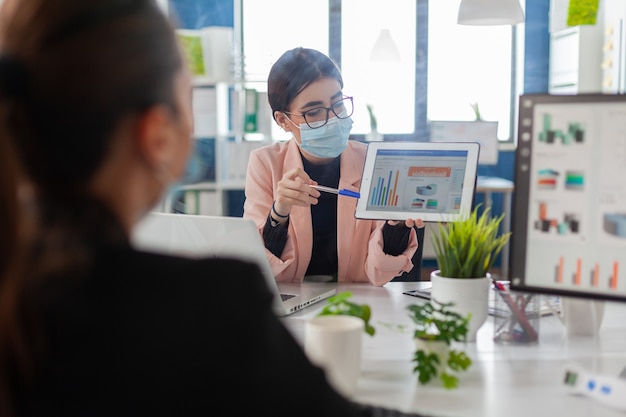 Nahaufnahme von Mitarbeitern mit Gesichtsmaske, die bei Finanzprojekten mit Tablet-Computer zusammenarbeiten, während sie im Firmenbüro sitzen. Das Team achtet auf soziale Distanz, um eine Ansteckung mit Covid19 zu vermeiden.