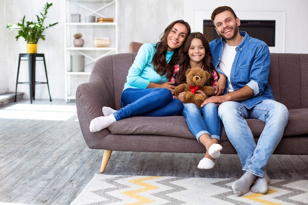 Nahaufnahme von Mutter, Vater und ihrer Tochter, die zusammen für ein gemeinsames Foto in ihrem stilvollen Wohnzimmer posieren und sich mit strahlendem Lächeln umarmen
