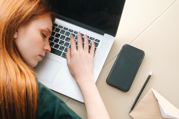 Nahaufnahme von oben einer überarbeiteten jungen Frau, die auf einer Laptop-Tastatur am Tisch mit einem Laptop im Heimbüro schläft. Erschöpfte Ermüdungsfreiberuflerin, die am Schreibtisch mit Computer an einem entfernten Arbeitsplatz schläft