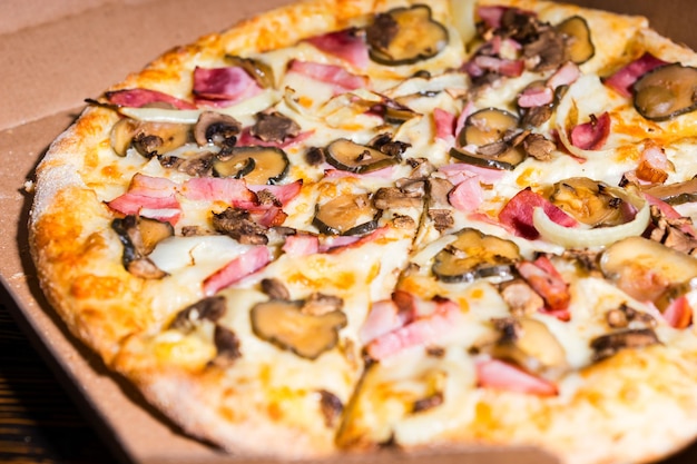 Nahaufnahme von Pizza mit Schinken, Champignons, Zwiebeln und Gurken in einer Kiste auf Holztisch