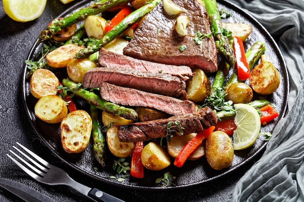Nahaufnahme von Rindersteak mit geröstetem Gemüse neue Babykartoffeln, Spargel und Paprika auf einem schwarzen Teller