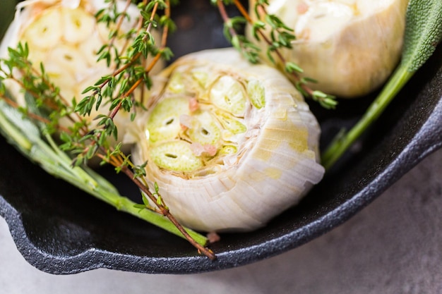 Nahaufnahme von rohen Knoblauchköpfen mit frischen Gewürzen in gusseiserner Pfanne zur Zubereitung von geröstetem Knoblauchrezept.