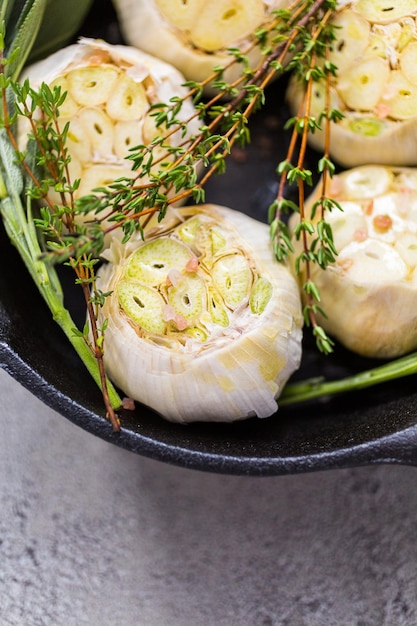 Nahaufnahme von rohen Knoblauchköpfen mit frischen Gewürzen in gusseiserner Pfanne zur Zubereitung von geröstetem Knoblauchrezept.