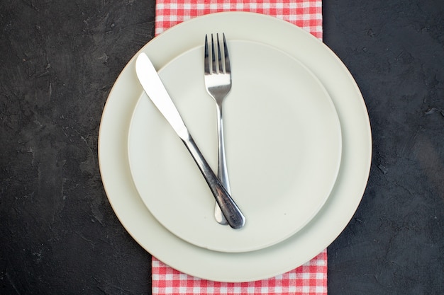 Nahaufnahme von rostfreiem Besteck auf weißen leeren Tellern auf rotem, abgestreiftem Handtuch auf schwarzem Hintergrund mit freiem Platz