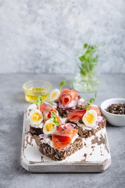 Nahaufnahme von Sandwich mit dunklem Roggenbrot, Frischkäse, Lachs, Zwiebeln, Kapern, gekochtem Ei auf weißem Holzbrett, Betonhintergrund. Traditionelles skandinavisches offenes Sandwich Smorrebrod