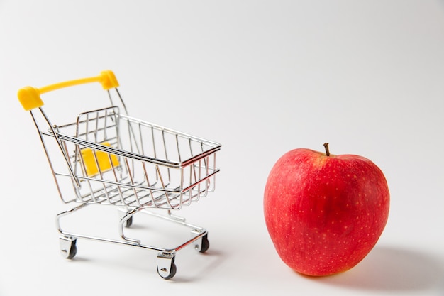 Nahaufnahme von Supermarkt Lebensmittel Push Cart zum Einkaufen mit gelben Kunststoffelementen am Griff in der Nähe von leuchtend rotem Apfel isoliert auf weißem Hintergrund. Konzept des Einkaufens. Kopieren Sie Platz für Werbung.