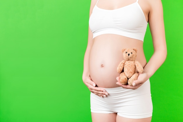 Nahaufnahme von Teddybären in der Hand der schwangeren Frau auf grünem Hintergrund. Die zukünftige Mutter trägt weiße Unterwäsche. Nackter Bauch. Konzept der Mutterschaft. Platz kopieren