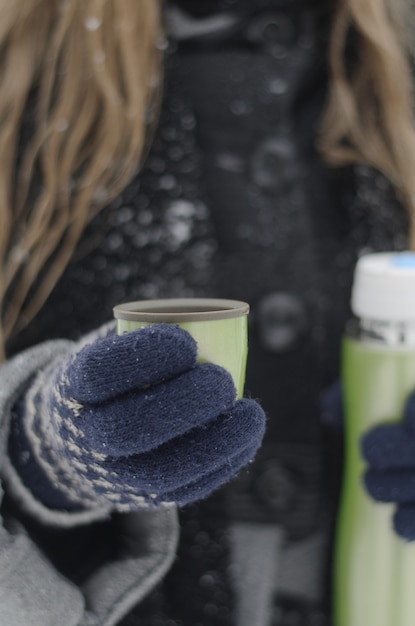 Nahaufnahme von weiblichen händen in gestrickten handschuhen, die heißen kaffee von einer thermoskanne an einem kalten winterschneetag gießen
