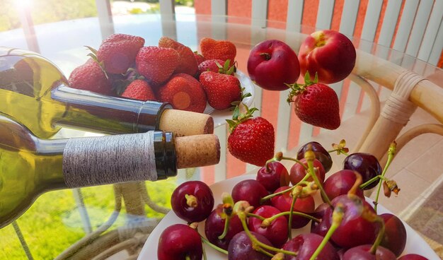 Nahaufnahme von zwei liegenden Weinflaschen, ein Haufen Früchte und Beeren auf einem transparenten Glastisch auf einem offenen Balkon an einem sonnigen Sommertag