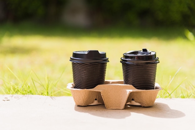 Nahaufnahme von zwei schwarzen Pappbechern heißen Kaffee zum Mitnehmen auf dem Boden vor dem Café auf dem Gartenplatz aus grünen Blättern Farbhintergrund für Kopierraum mit Sonnenlicht am Morgen