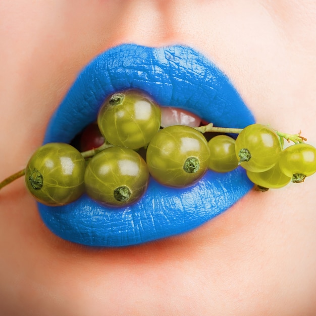 Nahaufnahme weißer Frauenmund mit blauen Lippen hält eine grüne Johannisbeere