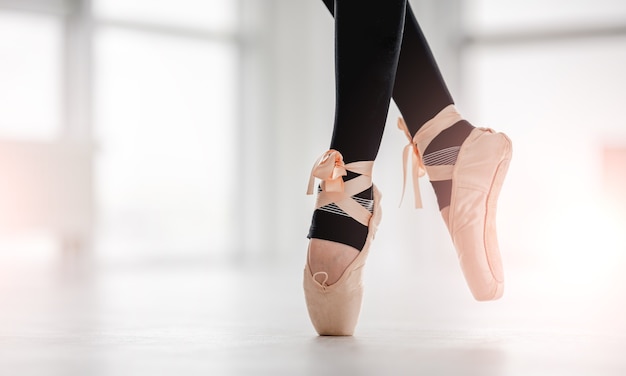 Nahaufnahmeansicht auf schönen Ballerina-Beinen, die schwarze Leggings und beige Spitzenschuhe tragen und auf Zehenspitzen im sonnigen Tanzstudio bleiben