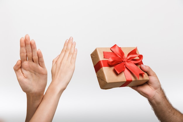 Nahaufnahmebild der Hände der Frau, die Stoppgeste zeigen, während ihr Mann über der weißen Wandwand lokalisiert wird, die gegenwärtige Geschenkbox hält.