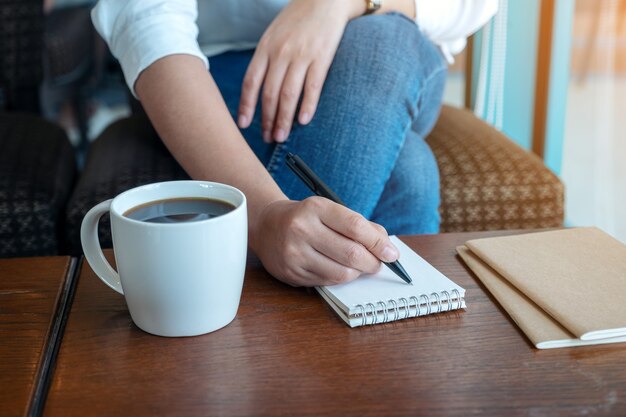 Nahaufnahmebild der Hand einer Frau, die auf leeres Notizbuch mit Kaffeetasse auf Holztisch schreibt