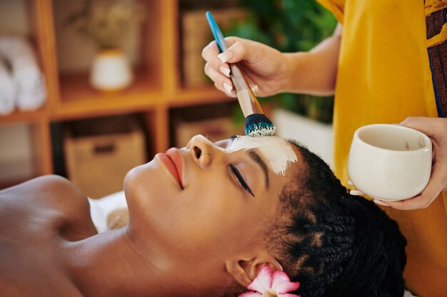 Nahaufnahmebild der Kosmetikerin, die reinigende und minimierende Poren-Tonmaske auf Gesicht der schönen jungen schwarzen Frau anwendet