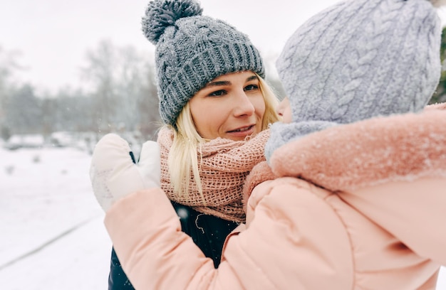 Nahaufnahmebild der schönen Mutter, die ihr Kind beim Gehen im Park an schneiendem Tag breit lächelnd hält Junge Frau und ihre Tochter, die zusammen spielen Weihnachtsstimmung Kindheit