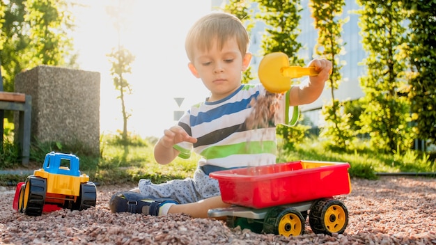 Nahaufnahmebild des netten kleinen Jungen, der auf dem Spielplatz mit Spielwaren spielt. Kind hat Spaß mit LKW, Bagger und Anhänger. Er gibt vor, ein Baumeister oder Fahrer zu sein