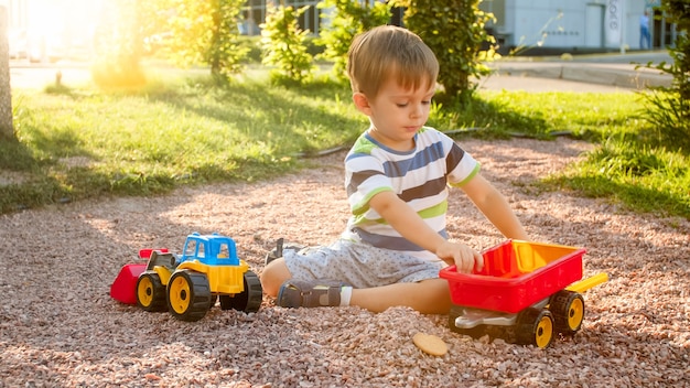 Nahaufnahmebild des netten kleinen Jungen, der auf dem Spielplatz mit Spielwaren spielt. Kind hat Spaß mit LKW, Bagger und Anhänger