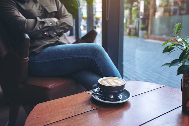 Nahaufnahmebild einer blauen Tasse heißen Lattekaffees auf Holztisch mit Frau, die im Café sitzt