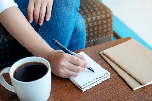 Nahaufnahmebild einer Frau, die auf leeres Notizbuch mit Kaffeetasse auf Holztisch schreibt