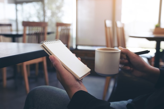 Nahaufnahmebild einer Frau, die ein leeres Notizbuch und eine Tasse heißen Kaffees beim Sitzen im Café hält