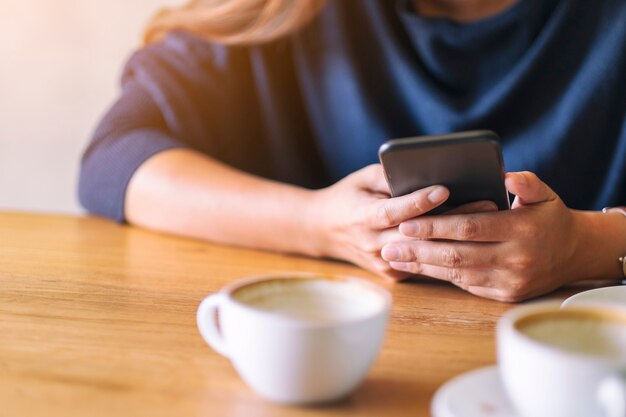 Nahaufnahmebild einer Frau, die Handy mit Kaffeetasse auf Holztisch hält und benutzt