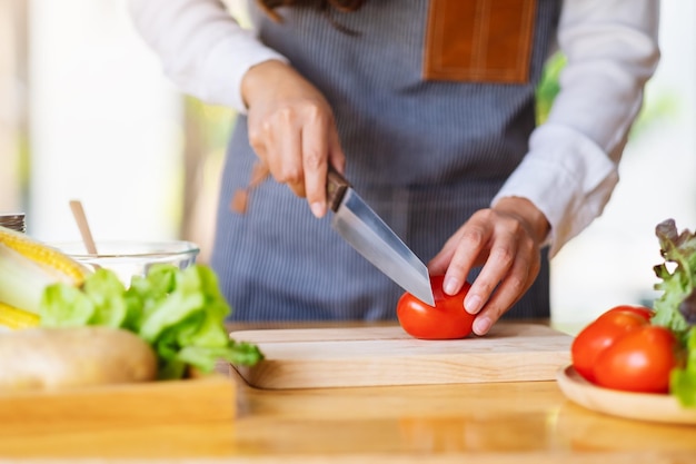 Nahaufnahmebild einer Frau, die Tomate mit dem Messer auf einem Holzbrett schneidet und hackt