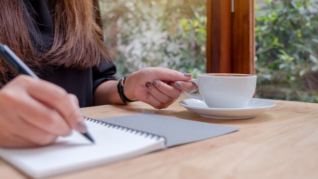 Nahaufnahmebild einer Hand der Frau, die auf ein weißes leeres Notizbuch schreibt, während Kaffee auf Holztisch trinkt