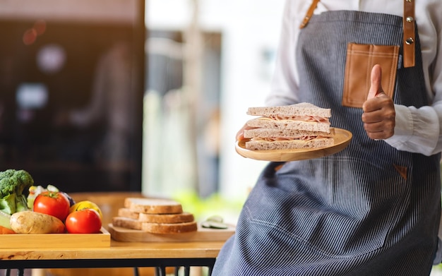 Nahaufnahmebild einer Köchin, die Daumen hoch Handzeichen macht, während sie einen Teller mit Vollkorn-Schinken-Käse-Sandwich in der Küche hält