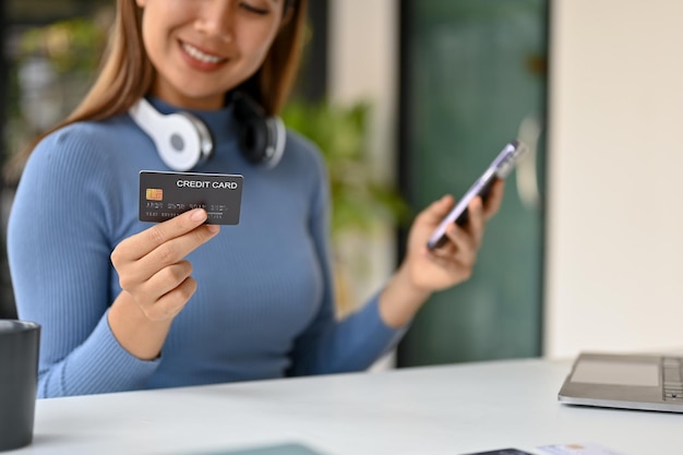 Nahaufnahmebild einer lächelnden asiatischen Frau sitzt an einem Tisch und hält ihre Kreditkarte und ihr Smartphone