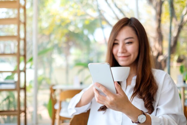 Nahaufnahmebild einer schönen asiatischen frau, die handy hält und benutzt, während sie kaffee im café trinkt