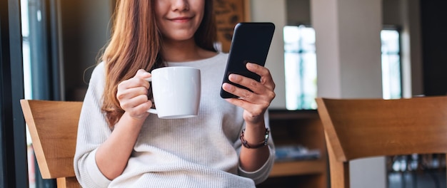 Nahaufnahmebild einer schönen asiatischen Frau, die Handy hält und verwendet, während sie Kaffee im Café trinkt