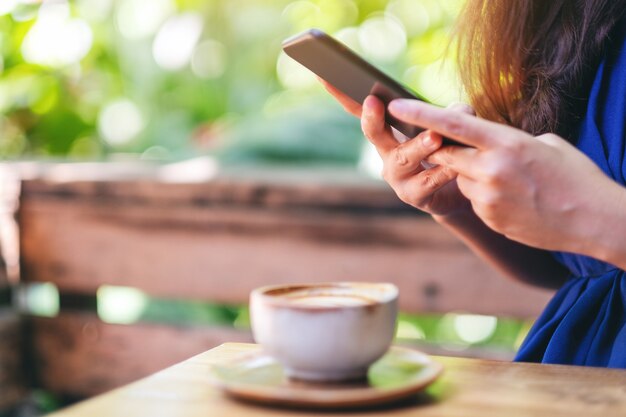 Nahaufnahmebild einer schönen asiatischen frau, die handy mit kaffeetasse auf dem tisch im garten hält und benutzt