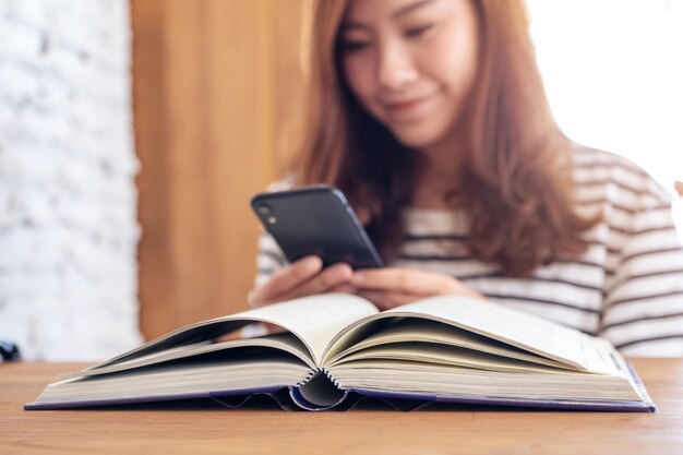 Nahaufnahmebild einer schönen asiatischen Frau, die Smartphone mit einem Buch auf Holztisch hält, benutzt und betrachtet