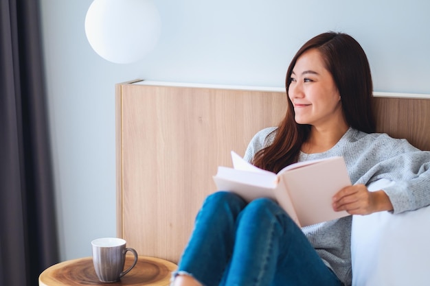 Nahaufnahmebild einer schönen jungen asiatischen frau, die buch in einem weißen gemütlichen bett zu hause liest