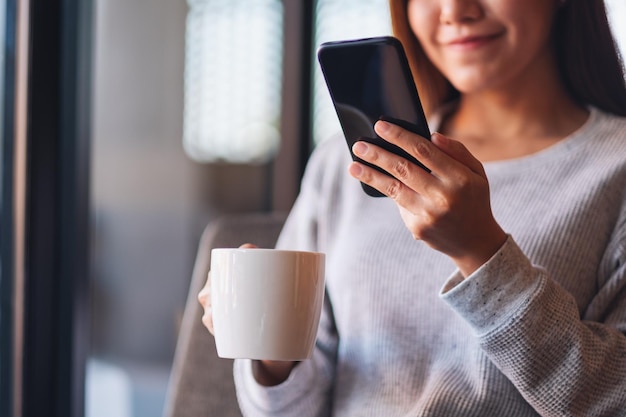 Nahaufnahmebild einer schönen jungen asiatischen Frau, die Handy hält und verwendet, während sie Kaffee trinkt