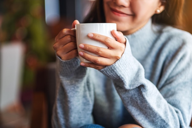 Nahaufnahmebild einer schönen jungen asiatischen Frau, die heißen Kaffee im Café hält und trinkt