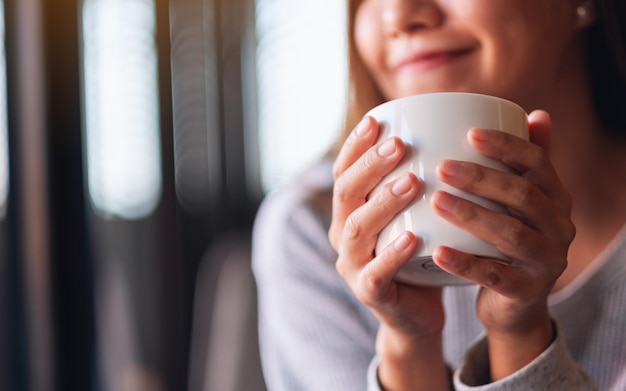 Nahaufnahmebild einer schönen jungen asiatischen Frau, die heißen Kaffee im Café hält und trinkt