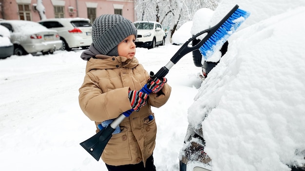 Nahaufnahmebild eines Jungen im beigefarbenen Mantel säubern das Auto nach Schneesturm mit Bürste