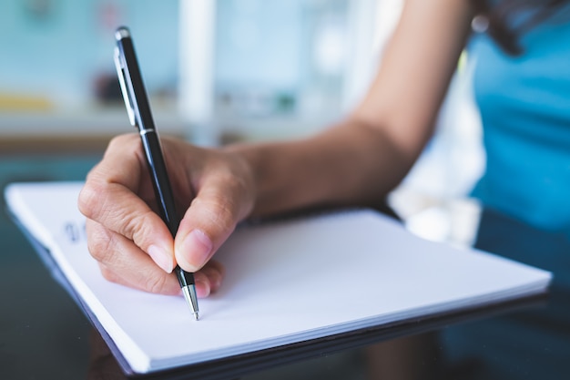 Nahaufnahmebilder von Frauen, die einen schwarzen Stift verwenden, um auf ein leeres Notizbuch auf einem Glastisch zu schreiben