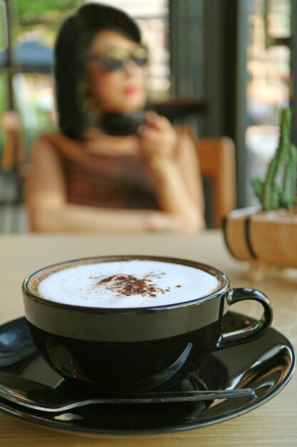 Nahaufnahmecappuccino in einer schwarzen tasse mit verschwommener frau, die kaffee im hintergrund trinkt