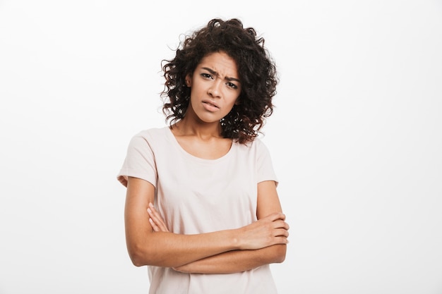 Nahaufnahmefoto der jungen afroamerikanischen frau 20s, die lässiges t-shirt mit stirnrunzeln hält, die arme verschränkt halten, lokalisiert über weißer wand